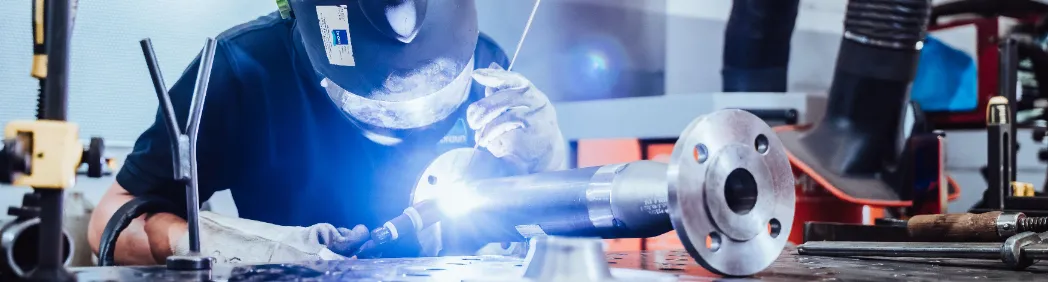 Schweißer bei der Arbeit in einer Werkstatt: Person mit Schutzmaske schweißt ein Metallrohr mit Flansch, Funken und helles Licht sichtbar.