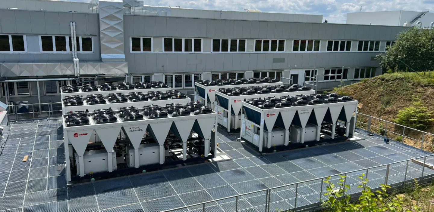 Außenansicht einer Industrieanlage mit mehreren großen Kälte- und Klimageräten der Marke Trane, aufgestellt auf einem Gitterboden vor einem modernen Gebäudekomplex unter blauem Himmel.