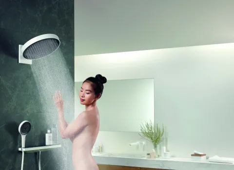Eine Frau mit hochgestecktem Haar steht unter einer modernen, runden Regendusche in einem minimalistisch gestalteten Badezimmer. 