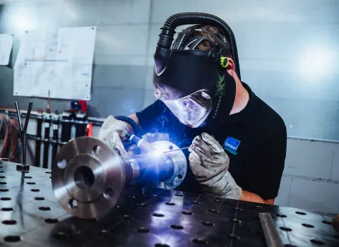 Schweißer mit Schutzhelm und Handschuhen bei präzisen Schweißarbeiten an einem Metallrohr auf der Werkbank, Funken leuchten auf.