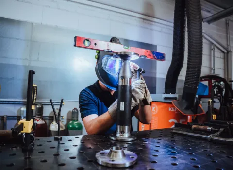 Schweißer mit Schutzhelm und Handschuhen bei der Arbeit an einem Metallrohr, ausgerichtet mit einer Wasserwaage auf der Werkbank.