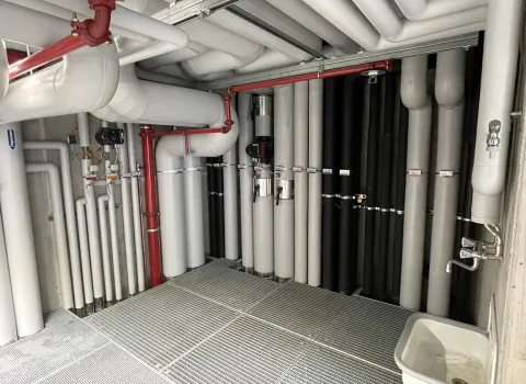 Technikraum mit zahlreichen isolierten Rohrleitungen für Abwasser- und Kälteanlagen, inklusive roten Sicherheitsleitungen und Waschbecken.