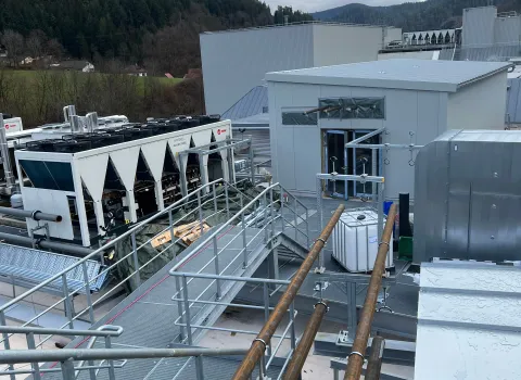 Dachinstallation einer Industrieanlage mit mehreren großen Kältemaschinen, Rohrleitungen und Wartungsstegen vor einer Berglandschaft.