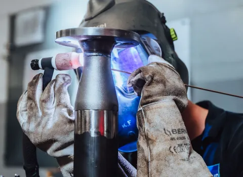 Handwerker mit Schutzhandschuhen und Schweißhelm beim präzisen Schweißen eines Metallrohrs.