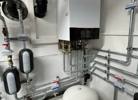 Moderne Heizungsanlage mit Wandgerät, Rohrleitungen und Ausgleichsbehälter in einem Technikraum.