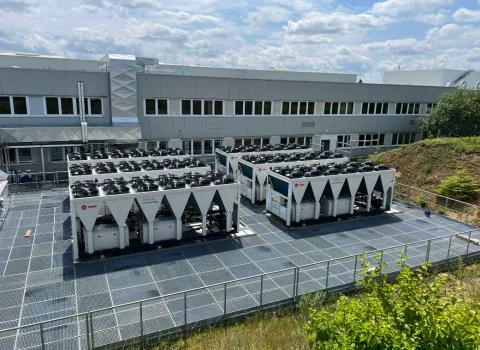 Außenansicht einer Industrieanlage mit mehreren großen Kälte- und Klimageräten der Marke Trane, aufgestellt auf einem Gitterboden vor einem modernen Gebäudekomplex unter blauem Himmel.
