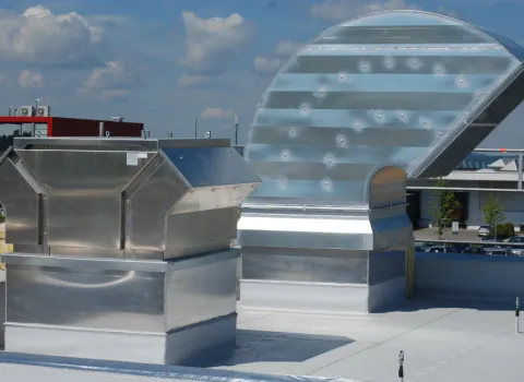 Dachansicht mit großen Lüftungs- und Klimaanlagen aus Metall: Luftkanäle und Abluftsysteme in glänzender Ausführung, im Hintergrund Industriegebäude und blauer Himmel.