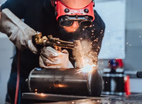 Handwerker mit Schutzhandschuhen und Schutzbrille beim Schweißen eines Metallzylinders, Funken sprühen während der Arbeit.