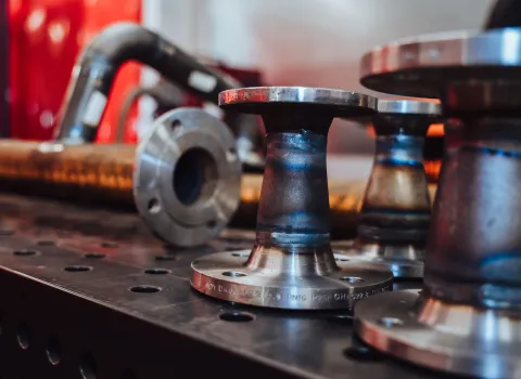 Frisch verschweißte Metallflansche auf einer Werkbank, im Hintergrund weitere Rohrstücke und Werkstattausrüstung.