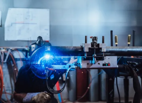 Schweißer mit Schutzhelm beim automatisierten Schweißvorgang an einem Rohr, blauer Lichtbogen und Funken sind deutlich sichtbar.