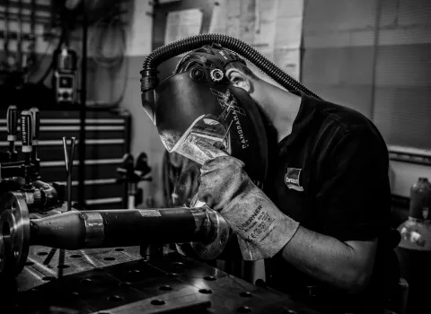 Schweißer mit Schutzhelm und Handschuhen bei präziser Metallbearbeitung an einem Rohr in der Werkstatt – Schwarz-Weiß-Aufnahme.