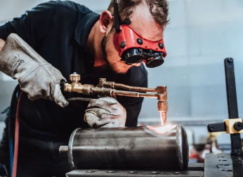 Handwerker mit Schutzhandschuhen und roter Schutzbrille beim Autogenschweißen eines Metallzylinders in der Werkstatt.