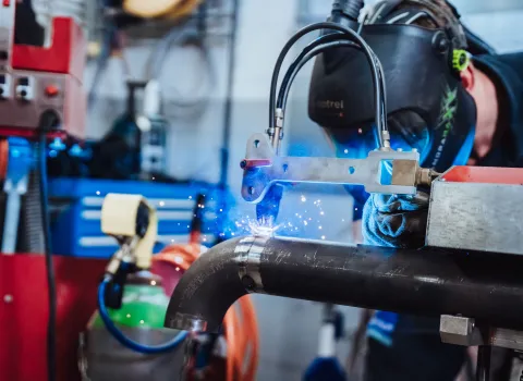 Schweißer mit Schutzhelm bei automatisiertem Schweißvorgang an einem Rohr, Funkenflug während der Arbeit in der Werkstatt.