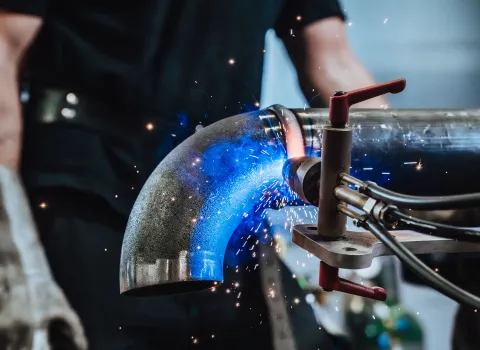 Nahaufnahme eines automatisierten Schweißvorgangs an einem Rohrbogen, blaue Flammen und Funken sprühen während der Arbeit.