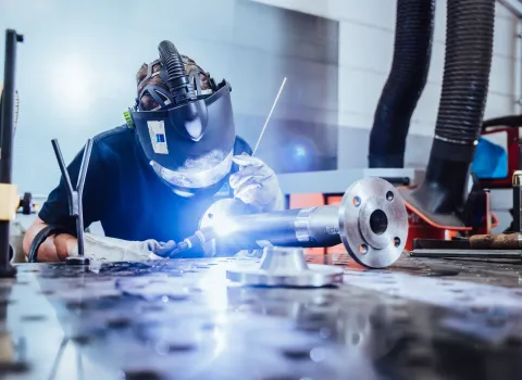 Schweißer mit Schutzhelm und Handschuhen bei präzisen Schweißarbeiten an einem Metallrohr auf der Werkbank.