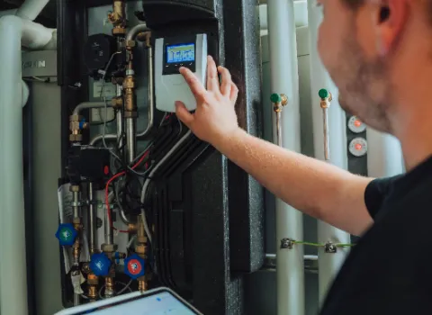 Servicetechniker prüft eine Heizungs- und Gebäudetechnik-Anlage: Bedienung des Steuerungsdisplays an der Anlage, Tablet mit digitaler Steuerungssoftware in der Hand.