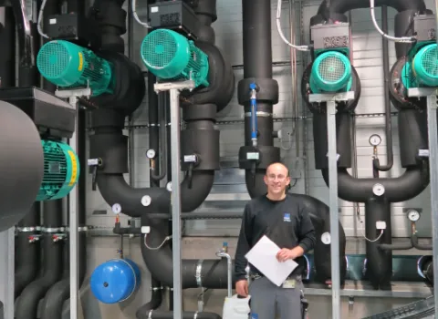 Techniker steht in einem modernen Technikraum mit großen Rohrleitungen, Pumpen und Steuerungssystemen, hält einen Plan in der Hand und lächelt in die Kamera.