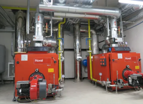 Technikraum mit zwei großen Heizkesseln der Marke Hoval, angeschlossene Rohrleitungen und Messinstrumente für die zentrale Heizungsanlage.