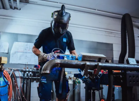 Schweißer mit Schutzhelm und Handschuhen führt einen Schweißvorgang an einem Rohrsystem in der Werkstatt durch.