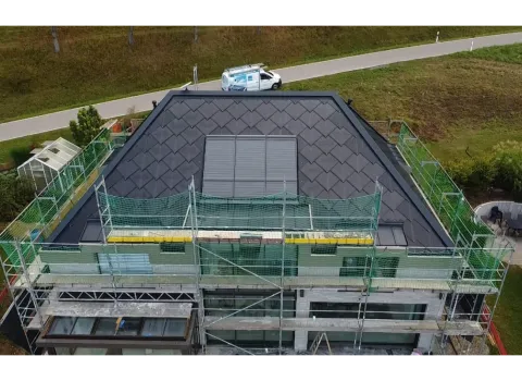 Luftaufnahme eines Hauses während der Dachsanierung. Das Dach ist bereits vollständig mit dunklen Schindeln gedeckt, in der Mitte befinden sich mehrere rechteckige Dachflächenfenster oder Solar-Module.