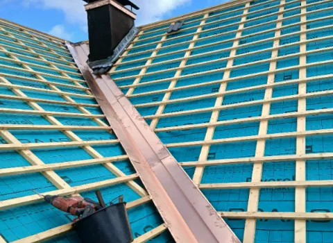 Dachaufnahme während der Sanierung: Eine Kupferkehle verläuft mittig zwischen zwei geneigten Dachflächen, die mit blauer Unterdeckbahn und einem Gitter aus hellen Holzlatten vorbereitet sind.