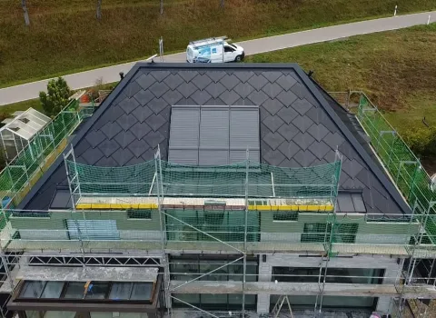 Luftaufnahme eines Hauses während der Dachsanierung. Das Dach ist bereits vollständig mit dunklen Schindeln gedeckt, in der Mitte befinden sich mehrere rechteckige Dachflächenfenster oder Solar-Module.