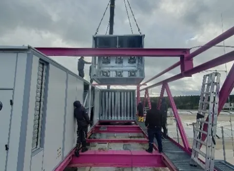 Ein großes technisches Modul wird mithilfe eines Krans auf eine pinkfarbene Stahlunterkonstruktion auf dem Dach eines Industriegebäudes gehoben.