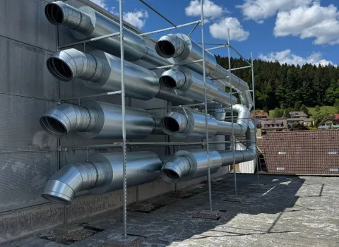 Mehrere große Lüftungsrohre aus glänzendem Metall sind ordentlich in einem Gerüst an der Außenwand eines Industriegebäudes montiert.