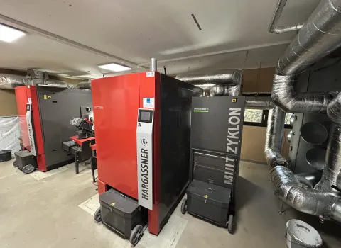 Ein Heizraum mit zwei leistungsstarken, rot-grauen Hackschnitzelheizkesseln der Marke Hargassner, verbunden mit einem großen Multizyklon-Filter zur Staubabscheidung.