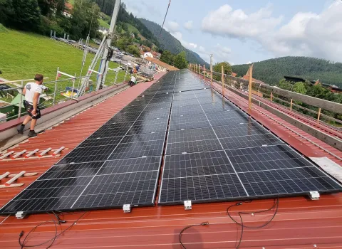 Montage von Solarmodulen auf einem roten Blechdach mithilfe eines Baukrans, umgeben von Wiesen und Hügeln.