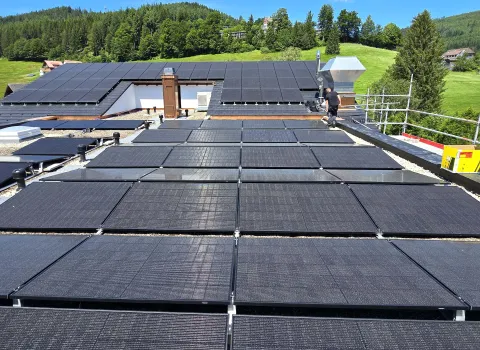 Montage von Solarmodulen auf einem roten Blechdach mithilfe eines Baukrans, umgeben von Wiesen und Hügeln.