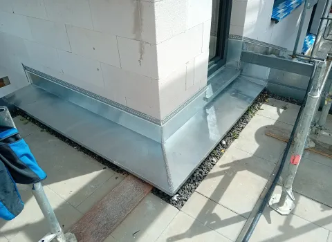 Metallene Anschlussbleche an einer Hausecke mit frisch montierter Fenster- und Sockelverblechung, umgeben von Pflasterflächen, Schotterbett und Gerüstteilen auf einer Baustelle.