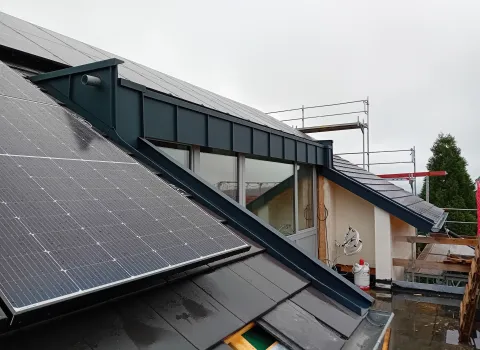 Dachansicht mit montierten Solarpanels neben einer metallverkleideten Gaube; darunter Fensterfront, umliegendes Gerüst und nasse Dachflächen auf einer Baustelle bei bewölktem Wetter.