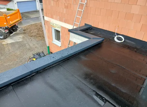 Frisch abgedichtetes Flachdach mit dunkler Bitumenoberfläche, umlaufender Metallabdeckung an der Hauswand, Bauleiter und Baustellenequipment im Hintergrund vor unverputztem Mauerwerk.