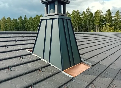 Modern verkleideter Dachaufbau mit Kaminaufsatz auf einem dunkel gedeckten Ziegeldach, umgeben von Wald und bewölktem Himmel, mit einem Baukran im Hintergrund.