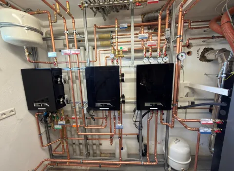 Das Bild zeigt eine hochmoderne Heizungssteuerung mit drei schwarzen ETA-Einheiten an einer weißen Wand, die über ein komplexes Netz aus Kupferrohren verbunden sind.