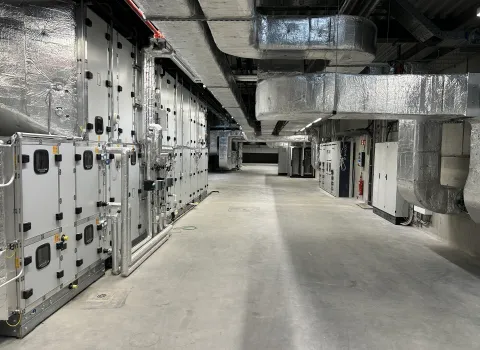 Ein moderner Maschinenraum mit großflächiger Lüftungsanlage zeigt zahlreiche silberne Luftkanäle und technische Anlagen entlang der Wand.