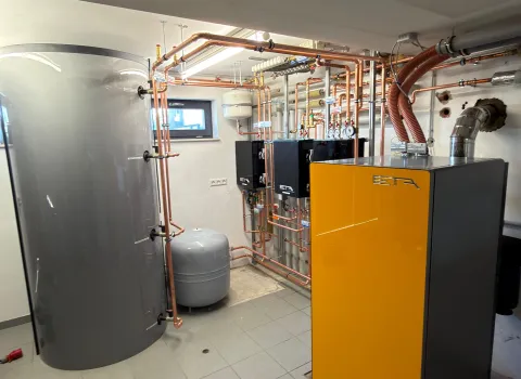 Das Bild zeigt einen hellen Heizungsraum mit einer modernen ETA-Pelletheizung in Gelb-Schwarz, einem großen grauen Pufferspeicher und zahlreichen Kupferrohren.