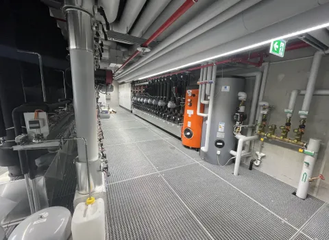 Das Bild zeigt einen modernen Technikraum mit umfangreicher Heizungs- und Regeltechnik, darunter ein großer Pufferspeicher, ein orangefarbener Wärmeerzeuger und zahlreiche Rohrleitungen.