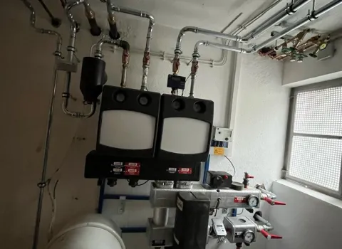 Ein kompakter Technikraum zeigt eine moderne Heizungsregelung mit zwei Wandmodulen, verbunden durch ein Netzwerk aus glänzenden Metallrohren.