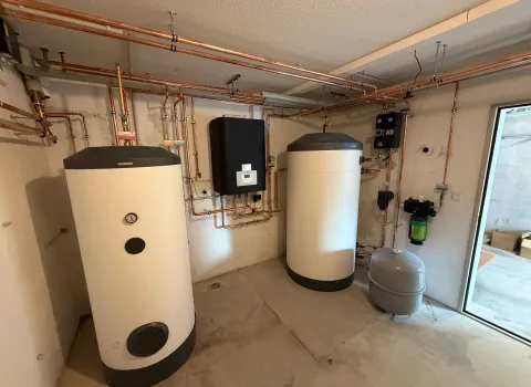 Ein Technikraum zeigt zwei große Warmwasserspeicher mit grauen Abdeckungen, die über ein komplexes Netzwerk aus neuen Kupferrohren verbunden sind.