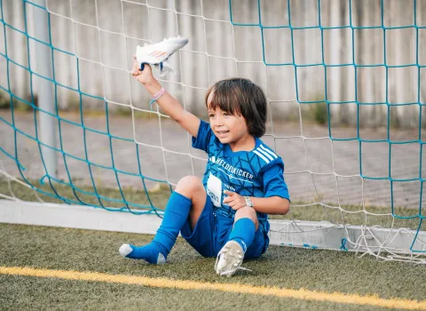 Ein Kind in blauen Heldenkicker-Trikot sitzt im Tor und hebt seinen verlorenen Fussballschuh in die Luft.