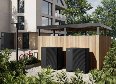 Zwei Waermepumpen-Außenheiten von Stiebel-Eltron stehen neben einem Mehrfamilienhaus vor einer Holzwand.