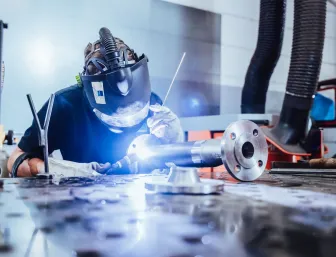Schweißer mit Schutzhelm und Handschuhen bei präzisen Schweißarbeiten an einem Metallrohr auf der Werkbank.