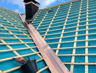 Dachaufnahme während der Sanierung: Eine Kupferkehle verläuft mittig zwischen zwei geneigten Dachflächen, die mit blauer Unterdeckbahn und einem Gitter aus hellen Holzlatten vorbereitet sind.