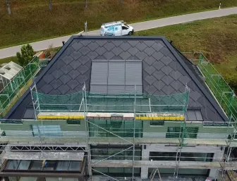 Luftaufnahme eines Hauses während der Dachsanierung. Das Dach ist bereits vollständig mit dunklen Schindeln gedeckt, in der Mitte befinden sich mehrere rechteckige Dachflächenfenster oder Solar-Module.