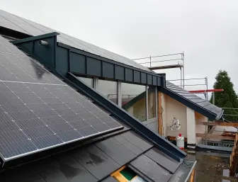 Dachansicht mit montierten Solarpanels neben einer metallverkleideten Gaube; darunter Fensterfront, umliegendes Gerüst und nasse Dachflächen auf einer Baustelle bei bewölktem Wetter.
