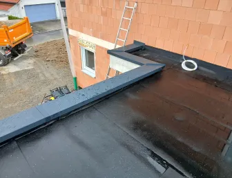 Frisch abgedichtetes Flachdach mit dunkler Bitumenoberfläche, umlaufender Metallabdeckung an der Hauswand, Bauleiter und Baustellenequipment im Hintergrund vor unverputztem Mauerwerk.