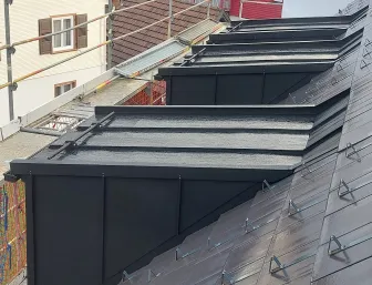 Mehrere dunkel verkleidete Dachgauben entlang eines geneigten Ziegeldachs mit Schneefanghaltern, eingerüstete Fassade im Vordergrund und umliegende Wohnhäuser im Hintergrund.