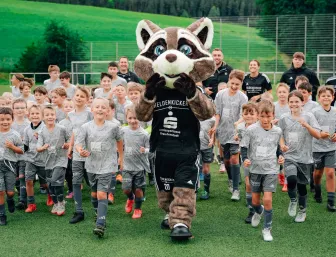 Vile Kinder im Fussballtrikot laufen hinter einem Maskottchen direkt Richtung Kamera.